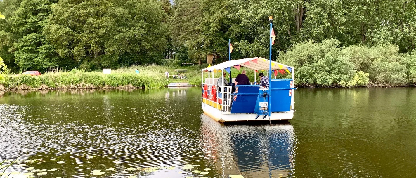 Emsquerung_mit_Passagiere_060622_Teipen.jpg Kleine Fähre überquert die Ems mit mehreren Passagieren, umgeben von grünem Ufer und Bäumen.