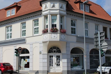 Een witte stadsvilla van twee verdiepingen met een rood pannendak en een decoratieve erker op de hoek van de straat.