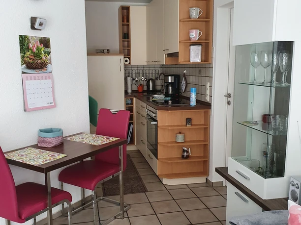 Küche & Esszimmer, Apartment Judt NEU Modern eingerichtete Küche mit hellem Holz, kleinem Esstisch mit roten Stühlen und Glasvitrine.