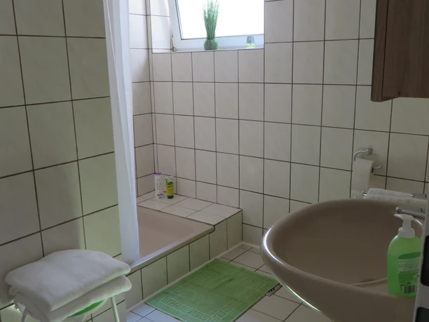Badezimmer - Apartment Judt Helles Badezimmer mit begehbarer Dusche, grünem Badvorleger und einem kleinen Fenster mit Pflanzen.