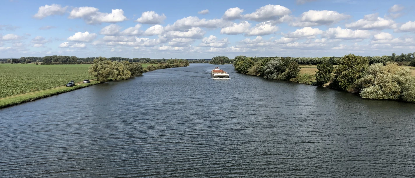 Breitflächiger Fluss, gesäumt von üppigem Grün, unter strahlend blauem Himmel mit ziehenden Wolken.