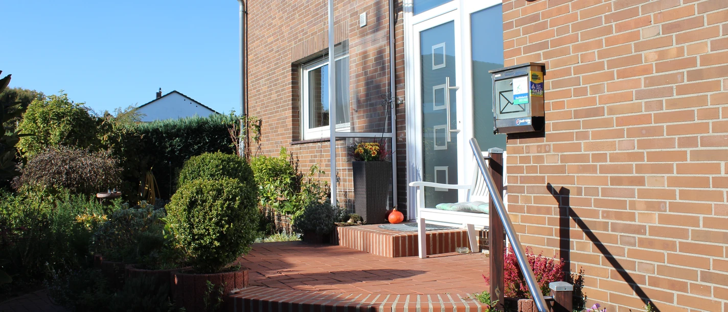 Entrance of a brick house with glass canopy and stairs, surrounded by plants and flowers.
