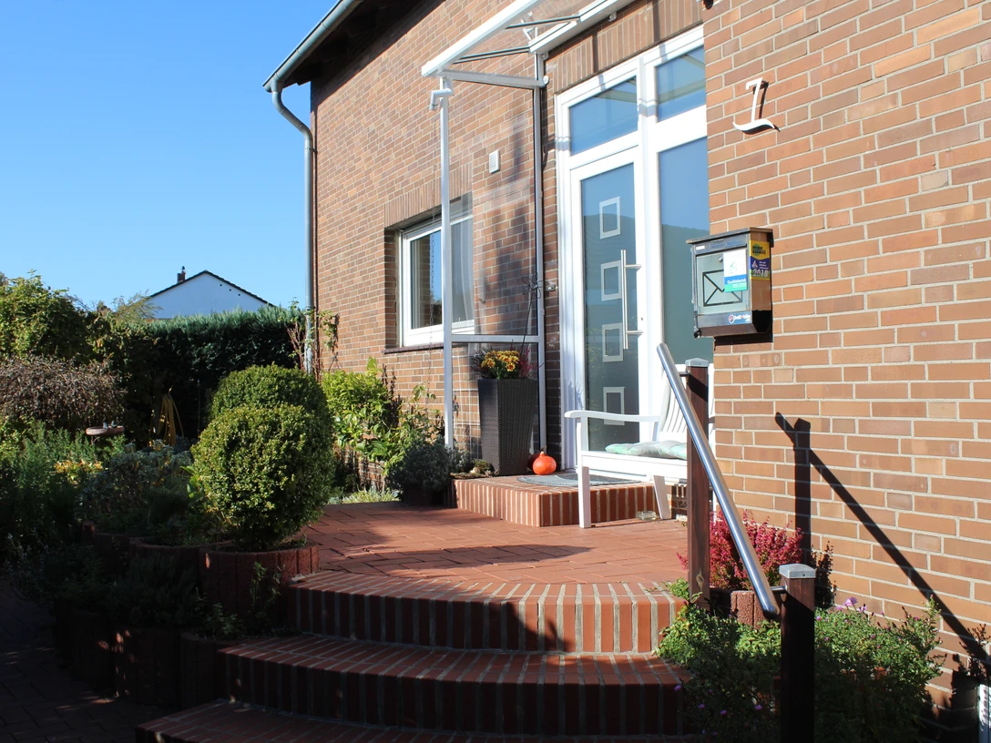 Frauenzimmer Eingang eines Backsteinhauses mit gläsernem Vordach und Treppe, umgeben von Pflanzen und Blumenschmuck.
