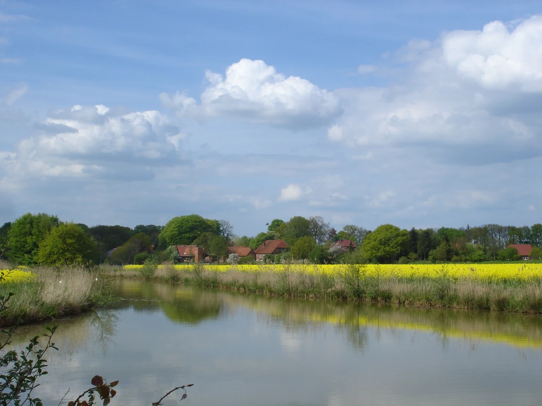 Gandesbergen1.JPG Landschaft mit Fluss im Vordergrund, gelbem Rapsfeld und Bauernhäusern unter bewölktem Himmel.
