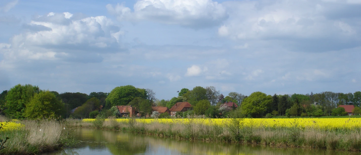 Gandesbergen1.JPG Landschaft mit Fluss im Vordergrund, gelbem Rapsfeld und Bauernhäusern unter bewölktem Himmel.