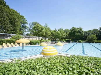 Aqualip Freibad Grünes Freibad mit großen gelben Pilzskulpturen, blauem Wasser und Bäumen umrahmt unter wolkenlosem Himmel.