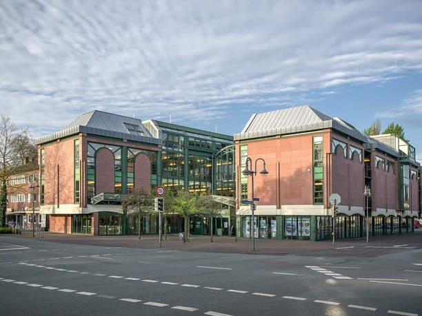Stadtbibliothek Gütersloh Großzügige Einkaufspassage mit roter Fassade und Glasfront, umgeben von Bäumen und einer Straße.