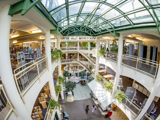 Eine lichtdurchflutete Bibliothek mit Glasdach, Pflanzen und offenen Büchergalerien auf mehreren Etagen.