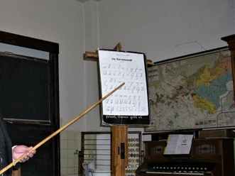 Schulmuseum Katterbach Ein Klassenzimmer mit einer Lehrtafel zur Kurrentschrift, einem Holzdreieck-Lineal und Landkarten an der Wand.