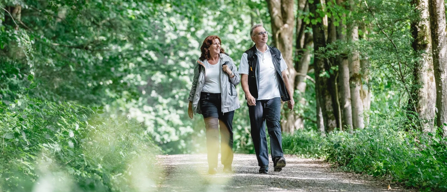 Zwei Personen spazieren auf einem Waldweg, umgeben von dichtem grünen Blattwerk.