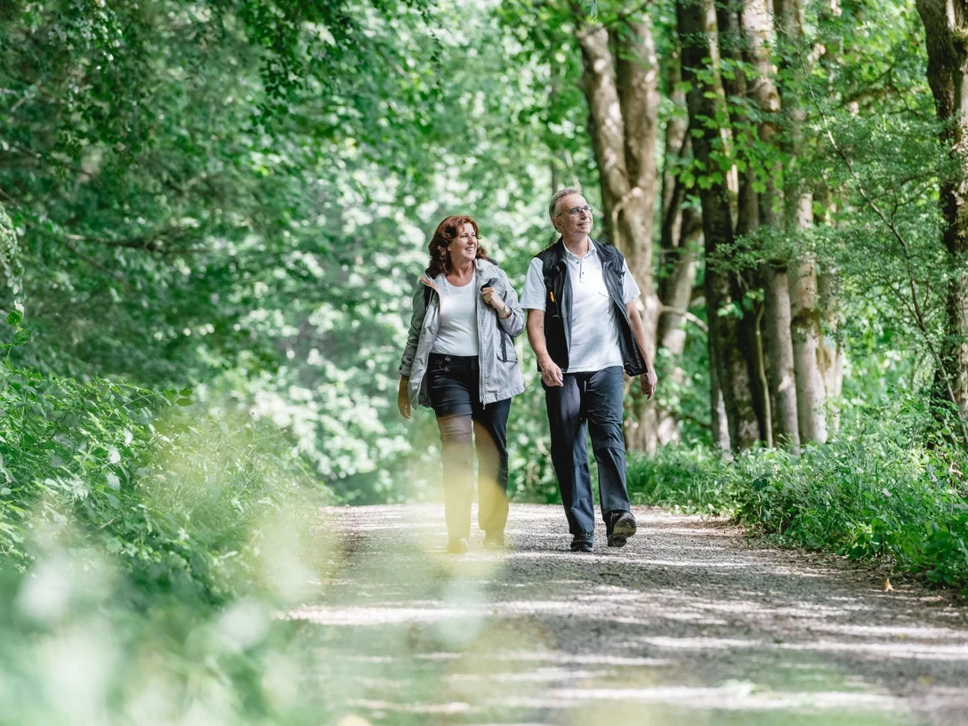 Geführte Wanderung Zwei Personen spazieren auf einem Waldweg, umgeben von dichtem grünen Blattwerk.