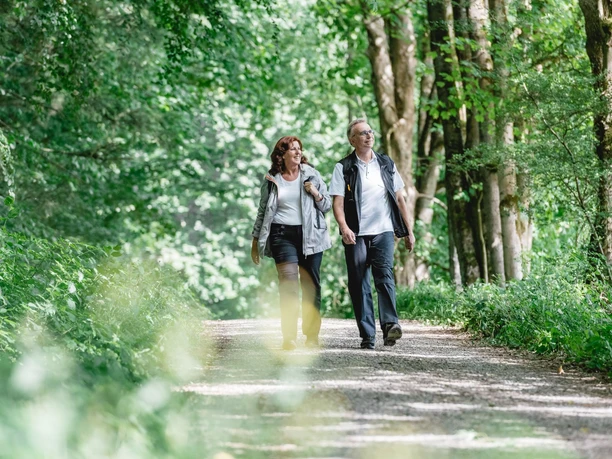 Geführte Wanderung Zwei Personen spazieren auf einem Waldweg, umgeben von dichtem grünen Blattwerk.
