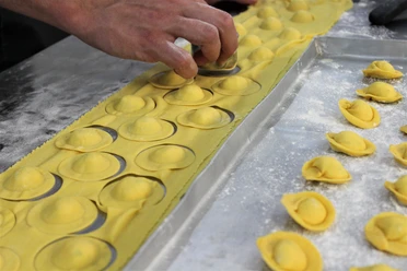 Eine Hand formt frische gelbe Pasta, während gefüllte Ravioli auf Mehl bestreutem Blech liegen.