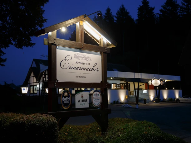 Restaurant Eimermacher Restaurant Eimermacher am Schloss bei Dämmerung, beleuchtetes Holzschild im Vordergrund.
