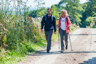 Geführte Wanderung Zwei fröhliche Wanderer spazieren auf einem sonnigen Weg durch die Natur, umgeben von grüner Vegetation.