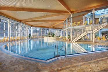 Toskana Therme Bad Schandau: helder binnenzwembad met blauw water, omgeven door gele ligstoelen en grote ramen; moderne, luchtige sfeer met houten plafond.