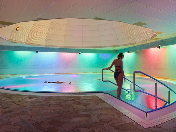 Toskana Therme Liquid-Sound Tempel Toskana Therme Bad Schandau: Eine Frau in einem schwarzen Badeanzug betritt ein Hallenbad mit bunten Lichtern; eine Person schwimmt im Wasser.