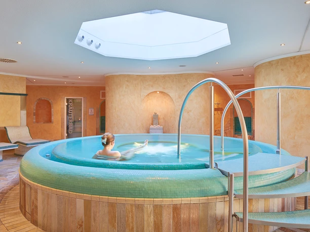 Toskana Therme Saunabereich Toskana Therme Bad Schandau: Eine Person entspannt in einem runden, türkisfarbenen Whirlpool in einem Spa mit warmen, beigen Wänden und einem großen Oberlicht.