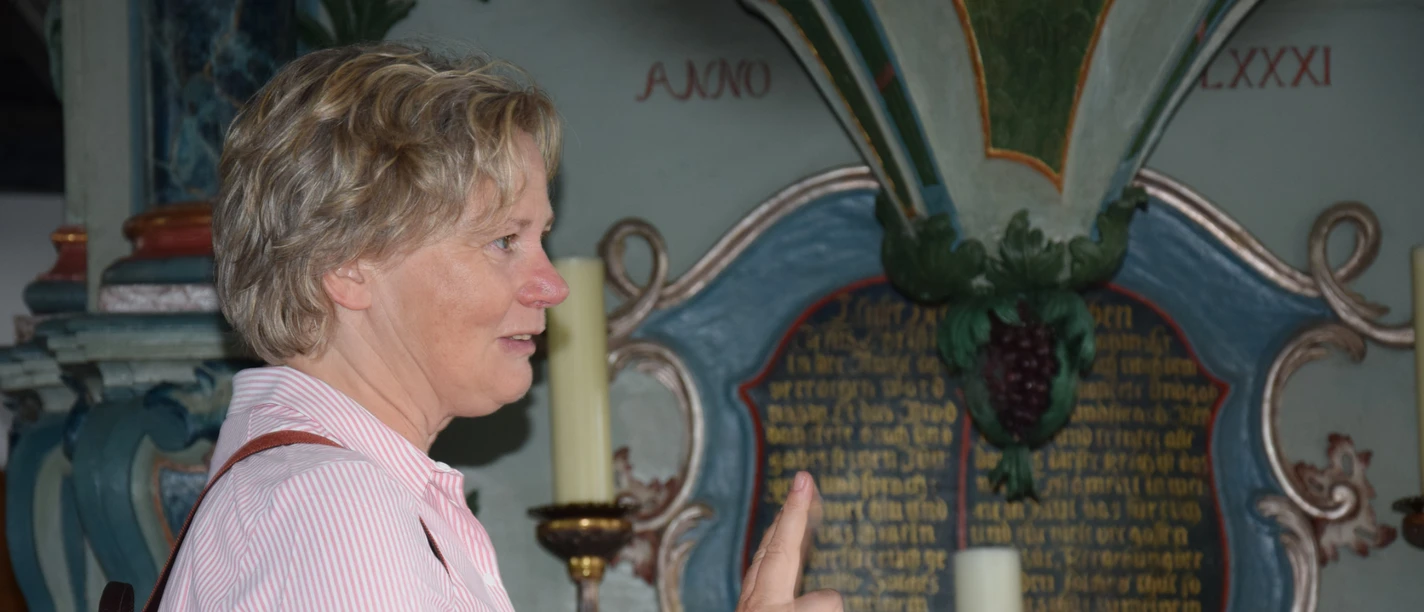 Regina Kerstin Frau in rotem Hemd nutzt Gebärdensprache in einem barocken Kirchenschiff, im Hintergrund historische Inschrift.