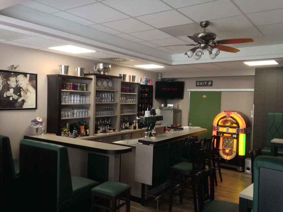 andreasklause.jpg Zu sehen ist die die Theke der Kneipe. Die Einrichtung der Kneipe ist in natürlichen Farben gehalten. In der Ecke si9ehrt man einen Spielautomaten.