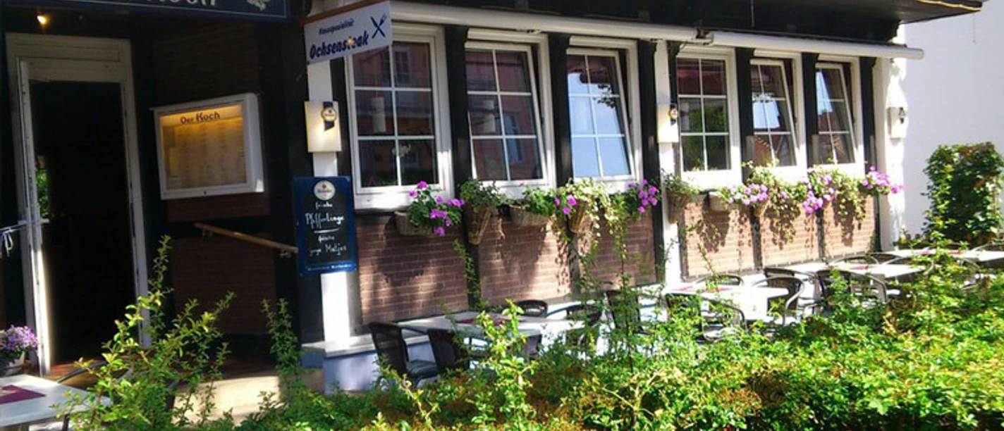 Restaurant Der Koch.jpg Auf dem Bild sieht man das Restaurant "Der Koch". Die Fassade des Gebäudes ist dunkel, mit weißen Fenstern, Blumenkästen und einer überdachten Terrasse.