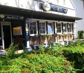 restaurant-derkoch.jpg Auf dem Bild sieht man das Restaurant "Der Koch". Die Fassade des Gebäudes ist dunkel, mit weißen Fenstern, Blumenkästen und einer überdachten Terrasse.