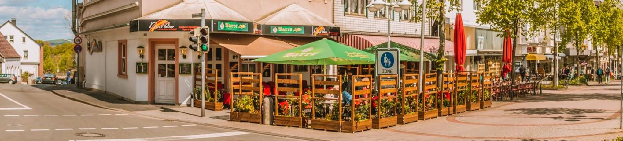 12232934_1017815684924633_8573930141605507645_o.jpg Zu sehen ist die Außenansicht des Restaurants Don Pedro. Die Fassade ist in weißen und braunen Farben gehalten. Vor dem Restaurant erkennt man mehrere Sitzgelegenheiten und zwei grüne Sonnenschirme.