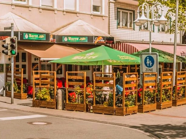 Restaurant Don Pedro Außenansicht.jpg Zu sehen ist die Außenansicht des Restaurants Don Pedro. Die Fassade ist in weißen und braunen Farben gehalten. Vor dem Restaurant erkennt man mehrere Sitzgelegenheiten und zwei grüne Sonnenschirme.