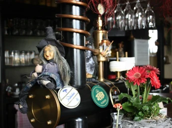 479859_579077668819427_933011793_n.jpg Das Bild zeigt eine Hexenpuppe im Vordergrund, die als Dekoration dienen soll. Neben ihr befindet sich ein kleiner Blumenstrauß. Im Hintergrund ist die Theke zu erkennen sowie weitere Weingläser.