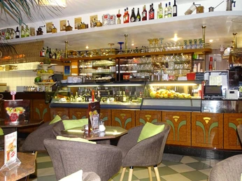 16112663_1444908805519555_502297902843371048_o.jpg Das Bild zeigt die gut bestückte Bar des Eiscafes. Im Vordergrund stehen mehrere Tische mit grünen Sitzpolstern, während im Hintergrund eine Theke mit Getränken, Gläsern und frischen Lebensmitteln zu sehen ist.