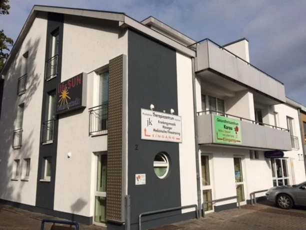 Appartement-Zimmer Karau.jpg Ein modernes, zweistöckiges Gebäude mit weißer Fassade, Balkonen und gewerblichen Räumen im Erdgeschoss.