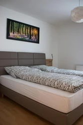 Appartement-Zimmer Karau_2.jpg Helles Schlafzimmer mit Doppelbett, grauem Kopfteil und Waldbild an weißer Wand, Holzboden.