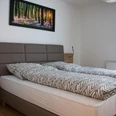 Appartement-Zimmer Karau_2.jpg Helles Schlafzimmer mit Doppelbett, grauem Kopfteil und Waldbild an weißer Wand, Holzboden.