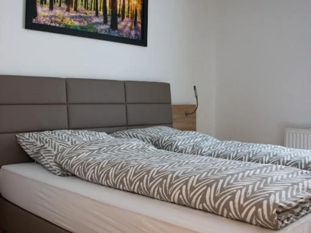 Appartement-Zimmer Karau_2.jpg Helles Schlafzimmer mit Doppelbett, grauem Kopfteil und Waldbild an weißer Wand, Holzboden.