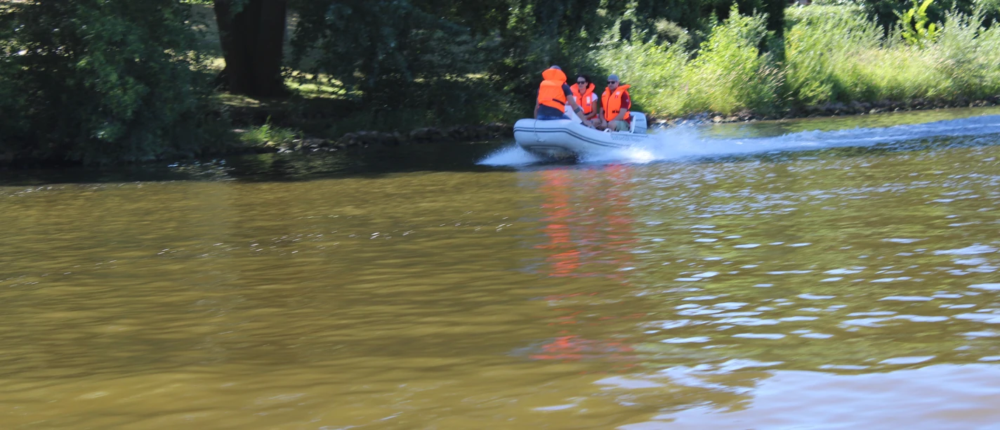 Bootstour_Weser_2018 (154).JPG Gruppe in einem Motorboot mit orangenen Westen fährt auf einem Fluss, umgeben von grünen Bäumen.