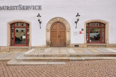 Witte gevel met bruine houten deur, geflankeerd door twee rode ramen met het opschrift "Tourist Service" en twee lantaarns aan de muur.
