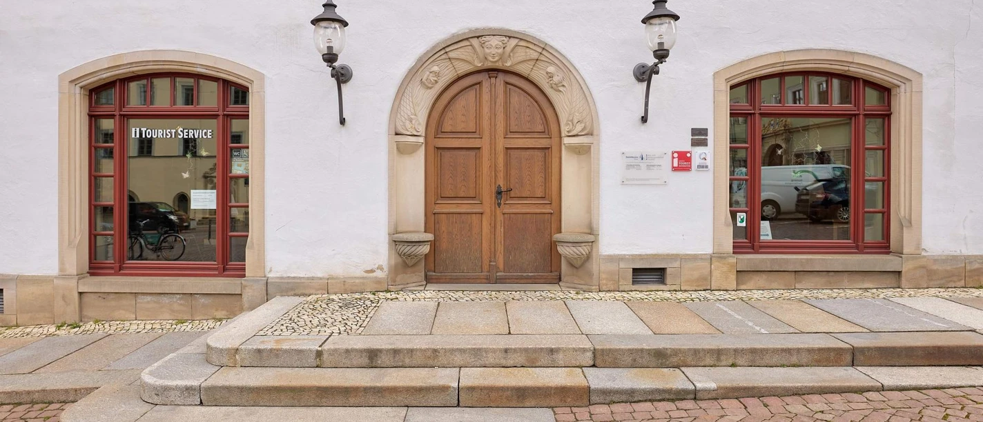 Canalettohaus Pirna Weiße Fassade mit brauner Holztür, flankiert von zwei roten Fenstern mit "Tourist Service"-Schriftzug, und zwei Laternen an der Wand.