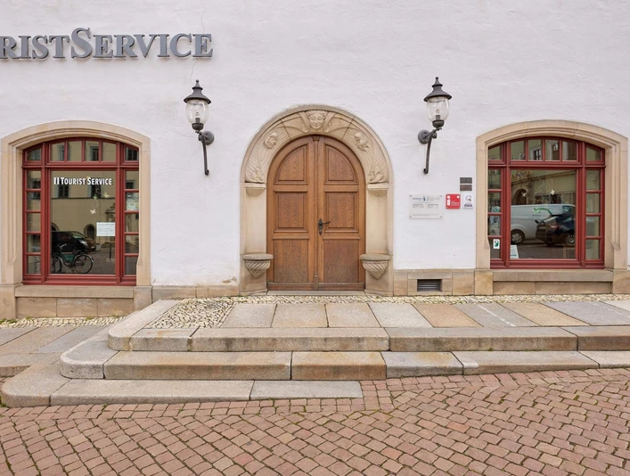 Canalettohaus Pirna Weiße Fassade mit brauner Holztür, flankiert von zwei roten Fenstern mit "Tourist Service"-Schriftzug, und zwei Laternen an der Wand.