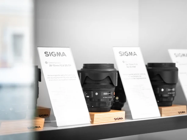 Drie Sigma cameralenzen op houten statieven op een plank, vergezeld van informatiekaarten. De sfeer is modern en minimalistisch.