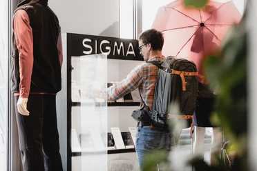 Een man met een rugzak bekijkt producten in een vitrine met het opschrift "SIGMA", terwijl er een etalagepop naast hem staat.