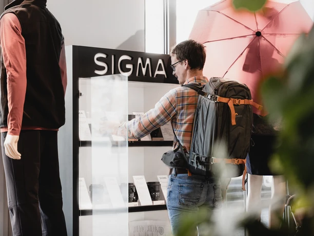 Een man met een rugzak bekijkt producten in een vitrine met het opschrift "SIGMA", terwijl er een etalagepop naast hem staat.