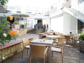 Buergerpark_Schaeppchen_1.jpg Heller Terrassenbereich mit Holztischen und Korbstühlen, umgeben von weißen Gebäudewänden.