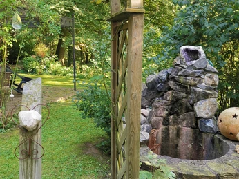 Waldkristall_2.jpg Ein hölzerner Gartenbogen vor einem Steinspringbrunnen, umgeben von Bäumen und Hecken im Garten.