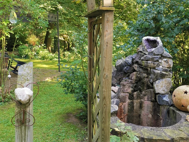 Waldkristall_2.jpg Ein hölzerner Gartenbogen vor einem Steinspringbrunnen, umgeben von Bäumen und Hecken im Garten.