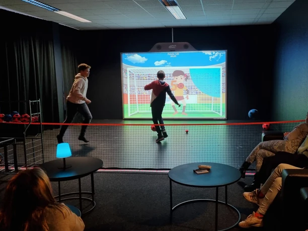 Dvě děti hrají pokutové kopy na XXL herní stěně Team-Duell Leipzig, zatímco diváci si dělají pohodlí v salonku a sledují zábavu při hře.