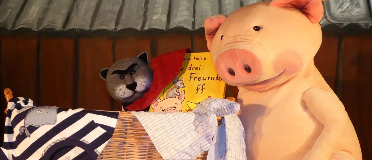 Ein großes Stoffschwein steht neben einem geflochtenen Korb mit gestreiften und karierten Stoffen.