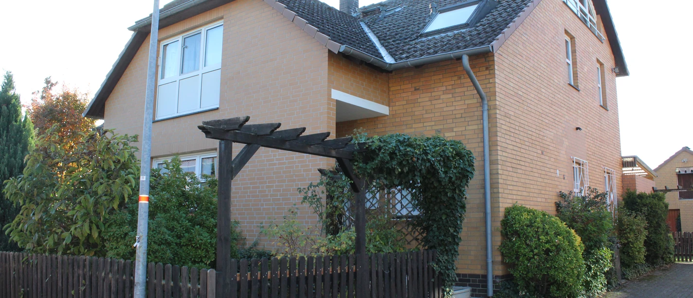 A two-storey brick house with a pitched roof, surrounded by hedges and a wooden fence in the foreground.