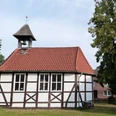 Historische Fachwerkkirche mit Glockenturm und rotem Ziegeldach, umgeben von grünem Rasen und Bäumen.