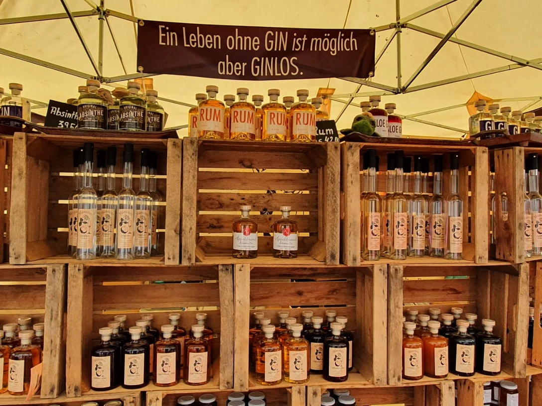 Handwerkermarkt: Gin Marktstand mit Regalen aus Holzkisten, gefüllt mit Gin-, Rum- und Absinthflaschen unter Zeltplane.