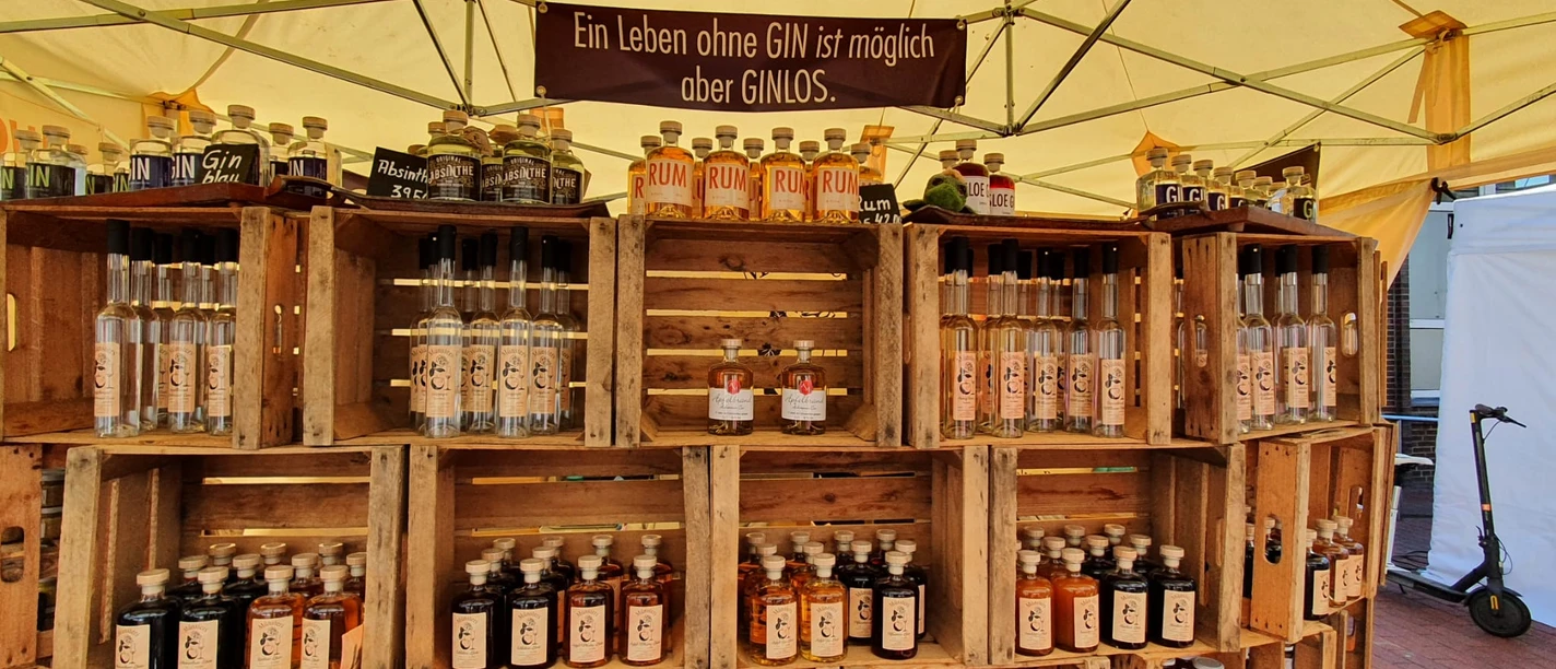 Handwerkermarkt: Gin Marktstand mit Regalen aus Holzkisten, gefüllt mit Gin-, Rum- und Absinthflaschen unter Zeltplane.
